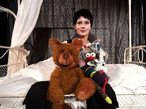  – Person mit kurzem dunklem Haar auf einem Bett hält Grossteddybaer und bunte Handpuppe mit weissen Haaren vor einem dunklen Vorhang.