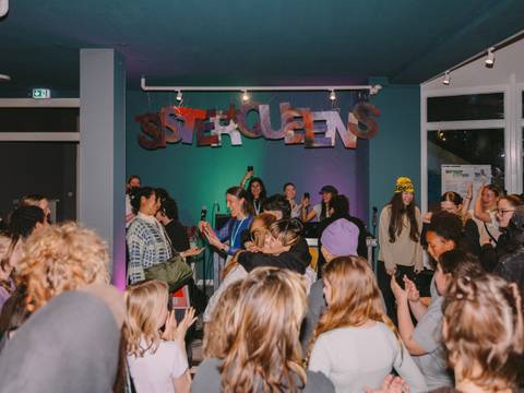  – Gruppe von Menschen in einem Raum, feiern unter einem bunten "SISTERQUEENS" Schild. Eine Person hält ein Mikrofon, andere klatschen und fotografieren.