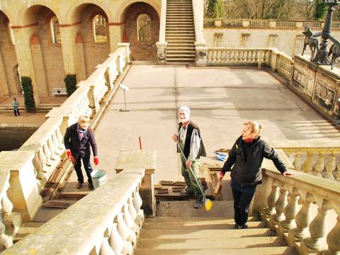 Putzen im Belvedere – Drei Personen reinigen eine Treppe mit Besen und Eimer in einem steinernen Innenhof mit Arkaden und Statuen.