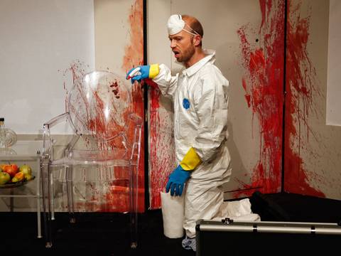  – Person in weissem Schutzanzug kniet vor roter Wand, hält Küchentuchrolle; durchsichtiger Stuhl und Obstschale links.