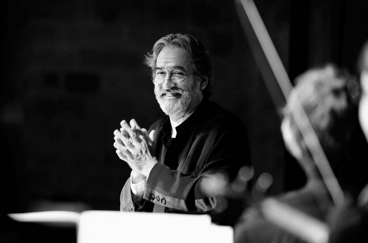 Jordi Savall – Ein älterer Mann mit Bart hält seine Hände vor der Brust verschränkt und lacht in die Kamera. Er steht vor einem Dirigentenpult an der Seite sind unscharf Streichinstrumente zu erkennen.