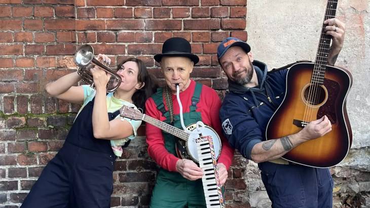 Die Drei von der Klangstelle – Drei Personen vor einer Backsteinmauer: links mit Trompete, in der Mitte mit Akkordeon und Hut, rechts mit Gitarre und Basecap.