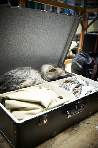Backstage-Führung – Offener Koffer mit grauhaariger Perücke, beigem Stoff, bunter Fliege und weitere Utensilien, im Hintergrund Holzgestell und Tasche.
