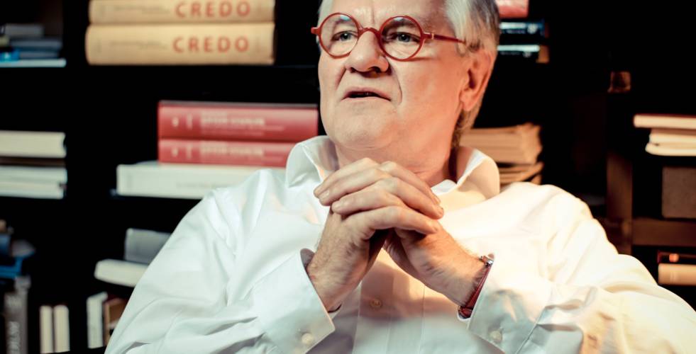 Ältere person mit roten Brillen, gefalteten Händen in weißem Hemd vor Bücherregal, Bücher mit Titel "CREDO" im Hintergrund.