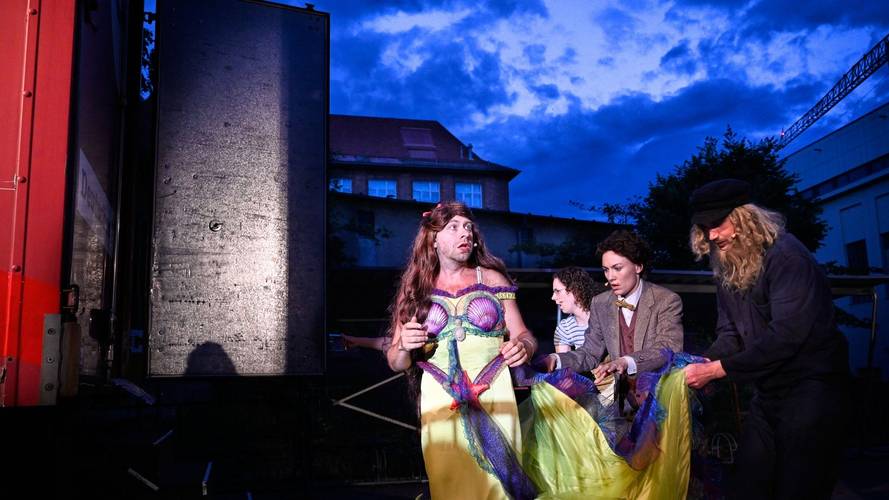 Vier Personen mit Kostümen im Freien, eine mit langem Haar und gelbem Kleid im Vordergrund, andere helfen. Gebäude und blauer Himmel im Hintergrund.