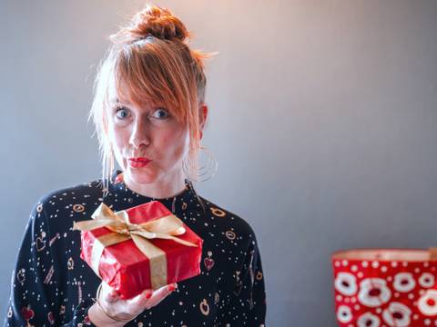 Wunschuniversum – Person mit Dutt hält ein rotes Geschenk mit gelber Schleife vor neutralem Hintergrund. Links im Hintergrund ein roter Beutel mit weißen Mustern.