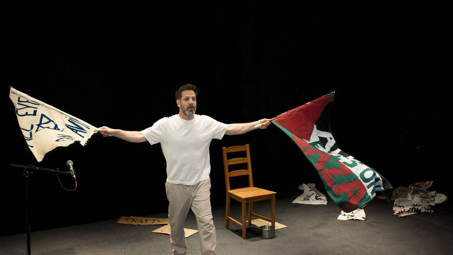 Between the River and the Sea – Person mit weißen T-Shirt hält zwei Fahnen; links blau-weiße Fahne, rechts rot-schwarz-grüne Fahne, Holzhocker und Mikrofonständer im Hintergrund.