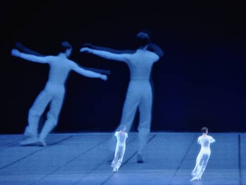  – Zwei Tänzer:innen auf einer leeren Bühne. Im Hintergrund eine Projektion der zwei Tänzer:innen.