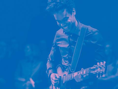 Key Visual Resident Music Collective – Lachende Person spielt E-Gitarre vor Publikum, blauer Farbstich.