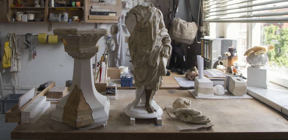 Frisch ergänzte Teilmodelle in der Werkstatt der Gipsformerei – Werkstatttisch mit einer kopflosen antiken Skulptur in Robe, einem Sockel und diversen Werkzeugen und Materialien im Hintergrund eines hellen Raums.