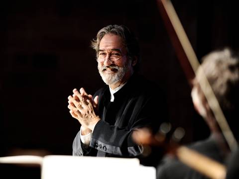 Jordi Savall – Männlich gelesene Person mit Brille und Bart, klatscht lächelnd in dunkler Kleidung. Im Vordergrund Instrumente unscharf.