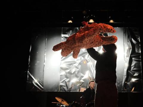  – Person hebt eine große, orange gepunktete Leopardenmarionette in die Luft. Hintergrund mit reflektierendem Material und Schlagzeug.