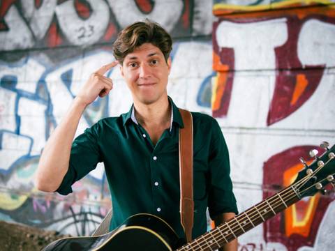 Falk - Unmöglich! – Person zeigt auf den Kopf, lächelt, hält akustische Gitarre; bunte Graffiti an der Wand im Hintergrund.