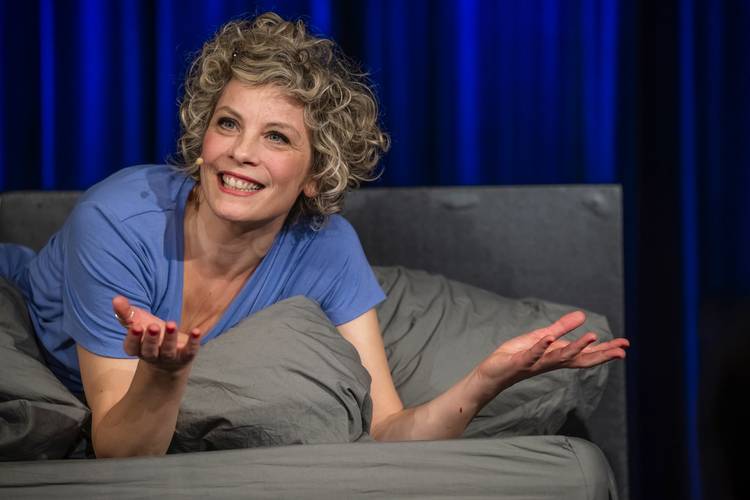 Person mit Locken und Mikrofon liegt auf grauem Bett, trägt blaues T-Shirt, breitet lachend die Arme aus, blauer Hintergrund.