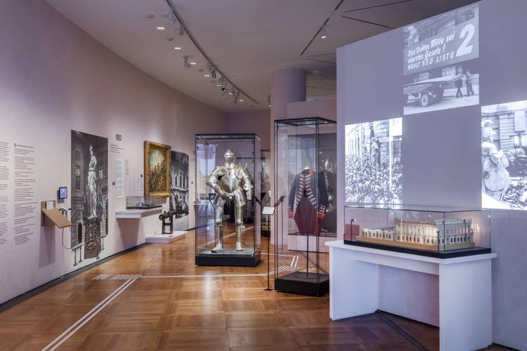 Raumansicht „Objekte. Geschichte. Geschichten. Blick in die Sammlung“. Im Vordergrund auf der rechten Seite ist eine Vitrine mit einem Modell des Zeughauses vor einer Projektionsfläche zu sehen. Auf dieser sind mehrere Schwarz-Weiß-Fotos zu erkennen. Im Hintergrund sind verschiedene Ausstellungsobjekte in Vitrinen zu sehen, darunter eine Ritterrüstung und eine Militäruniform. An der Wand auf der linken Seite hängen Gemälde und Fotos.