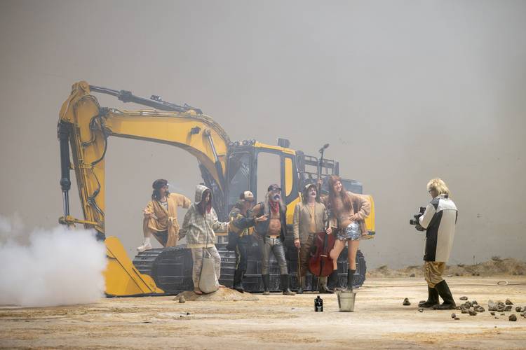 Gruppe von sechs Personen vor gelbem Bagger, eine mit Kamera, andere posieren mit Instrument; Umgebung rauchig, Boden steinig.