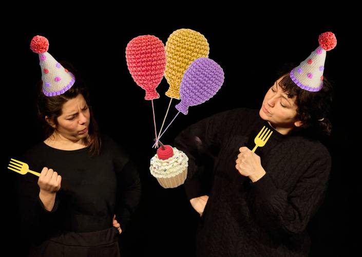 Zwei Personen mit Partyhüten, halten Plastigabeln, betrachten einen gehäkelten Cupcake und Luftballons vor schwarzem Hintergrund.