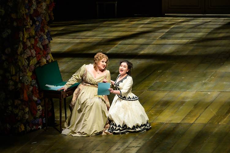 Le nozze di Figaro – Zwei Personen in Opernkostümen auf einer Bühne; eine sitzt an einem Tisch mit grünem Ordner, die andere kniet daneben. Hintergrund aus Blumen.