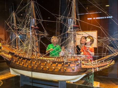 Die prachtvollen Schnitzereien der schwedischen VASA von 1628 zeigen, dass Schiffe auch als Repräsentationsobjekte und Ausdruck von Macht dienten. – Drei Kinder stehen auf einem Podest hinter einer Vitrine und betrachten ein großes, detailreiches Schiffsmodell. Hinter ihnen steht eine Frau.