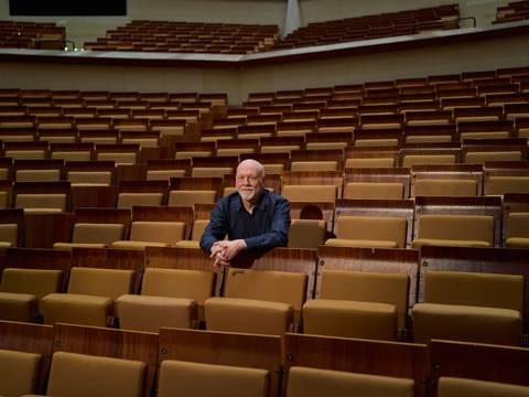 Brett Dean – Brett Dean sitzt allein in der Mitte eines großen, leeren Saals mit braunen Sitzreihen und blickt nach vorne, die Arme auf dem Sitz vor ihm gestützt.