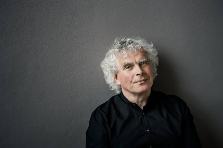 Sir Simon Rattle – Eine Person mit lockigem, grauem Haar trägt ein schwarzes Hemd und blickt leicht lächelnd in die Kamera vor grauem Hintergrund.