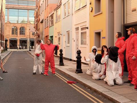  – Eine Gruppe von Personen steht und kniet in einer schmalen Stadtstraße zwischen farbigen Hausfassaden. Einige tragen rote Schutzanzüge, andere weiße Overalls mit Kapuzen; zwei knien am Boden mit erhobenen Händen, eine Person hat einen schwarzen Stoff über dem Kopf. Mehrere Beteiligte tragen Kopfhörer. Auf der linken Seite beobachten zwei Personen in Alltagskleidung die Szene. Die Situation wirkt wie eine inszenierte Performance im öffentlichen Raum.