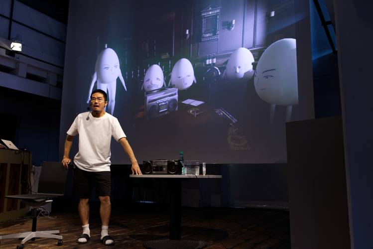 Person mit Mikrofon steht vor einer Projektion mit animierten Figuren. Tisch mit Flasche und Lautsprechern.