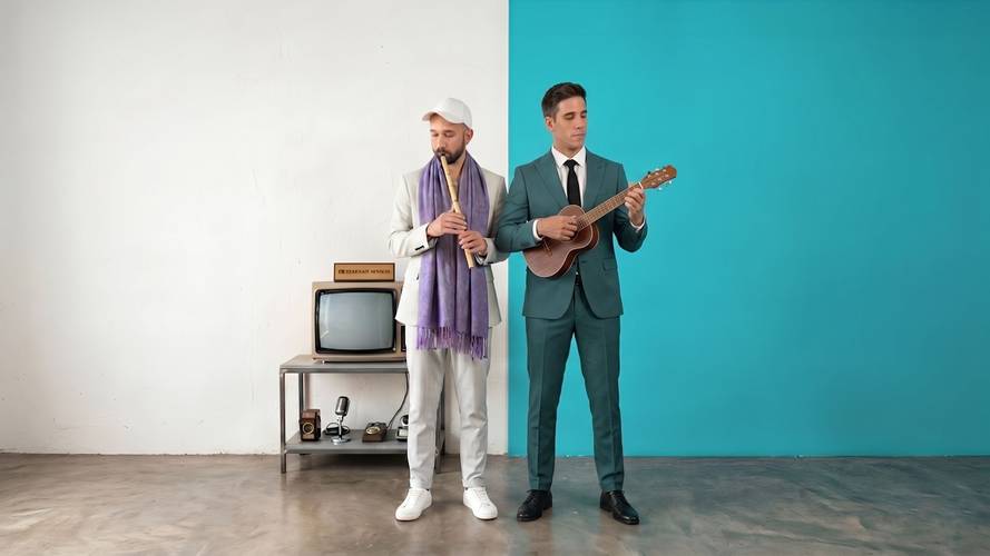 Zwei Personen: links weiß gekleidete Person mit Schal und Flöte, rechts Person im Anzug mit Gitarre. Im Hintergrund ein Fernseher und Mikrofone.