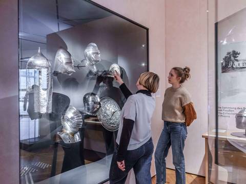  – Zwei Besucherinnen stehen vor einer Vitrine. Diese ist an der Wand befestigt. In der Vitrine sind mehrere Helme aus dem Mittelalter zu sehen. Die Besucherinnen beobachten sie.