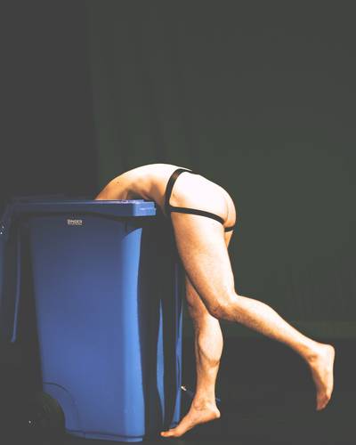 Performer:in in schwarzem Harness beugt sich auf einer dunklen Bühne über eine blaue Mülltonne.