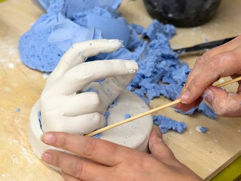  – Zwei Haende bearbeiten eine weiße Gips-Handform auf Holzplatte, umgeben von blauen Silikonbruchstuecken und einem Werkzeug.