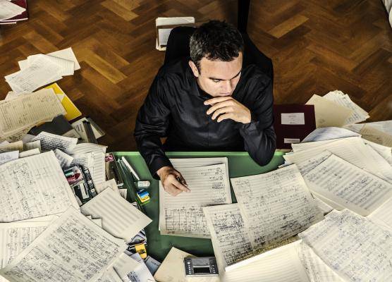 Jörg Widmann – Person am Schreibtisch, umgeben von einem Chaos aus Notenblättern und Papier, hält einen Stift in der Hand und wirkt nachdenklich, Holzboden im Hintergrund.