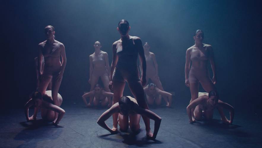 DART Dance Company: KOMPÓT – Personengruppe in Hautfarben-clothing auf Buhne. Zwei stehen, andere knien davor mit Kopf gesenkt. Lichtstrahl von oben beleuchtet Szene dramatisch.