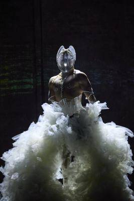 Queer Performance Festival – Person in dunkler Umgebung, trägt aufwendiges weißes Kostüm mit Federn oder Tüll und glitzernde Maske mit Ohren ähnlichem Kopfschmuck.