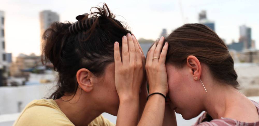 Choreographies of Listening – Zwei Personen stehen Kopf an Kopf, Hände bedecken ihre Gesichter. Hintergrund unscharf mit Hochhäusern. Fotografie.