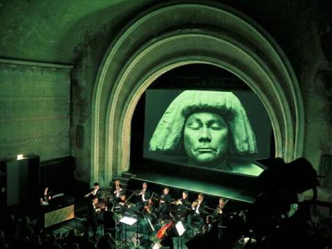 GOLEM – Orchester spielt in einem Theater mit hohem Bogen; große Leinwand zeigt Skulptur mit Augen geschlossen, Stirnband und langen Haaren.