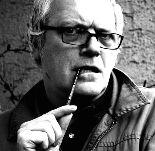 Porträt Wolf Hogekamp, Begründer und Erfinder der Deutschen Poetry-Slam-Meisterschaften – Mann mit grauen Haaren und Brille hält einen Stift im Mund, schaut bedacht und ernst in die Kamera. Schwarz-weiße Fotografie.