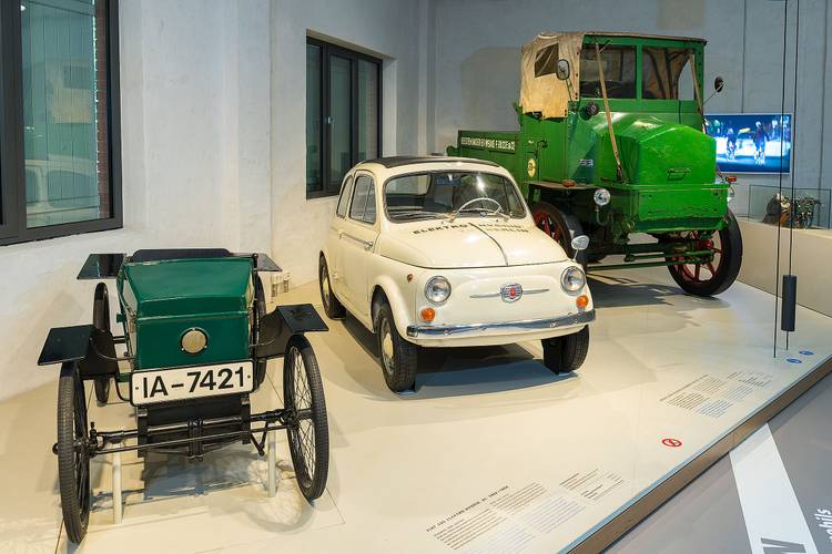 Schon zu Beginn des 20. Jahrhunderts wurden Elektro-Fahrzeuge gebaut. Die von AEG und Siemens produzierten Akkumulatoren, waren jedoch noch nicht ausgereift und sehr schwer. – Auf einem niedrigen Podest stehen drei historische Elektro-Fahrzeuge.Von links nach rechts: ein grüner offener Wagen, ähnlich einer Seifenkiste, ein weißer Fiat 500 und ein grüner LKW mit offener Ladefläche.