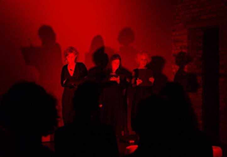 Drei Personen in schwarz in rotem Licht auf einer Bühne mit Backsteinwand, Schatten hinter ihnen. Publikum im Vordergrund als Silhouetten sichtbar.