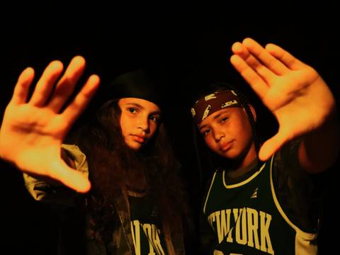 LA PREMIERE FOIS – Zwei Personen in Basketballtrikots mit dunkelgrünem „New York“-Schriftzug halten ihre Hände nach vorne, dunkler Hintergrund.