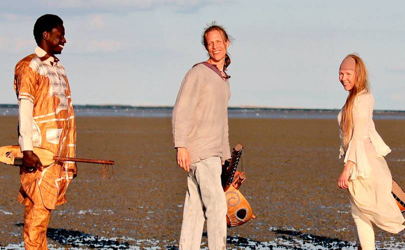 Drei Personen stehen am Strand, jede hält ein Saiteninstrument, lächelnd, mit dem Meer im Hintergrund.