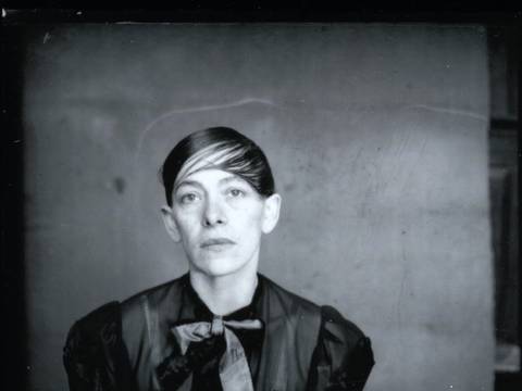 HeWe_Pressefoto_04.jpg – Person in schwarzer Kleidung mit großer Schleife, kurzes Haar zur Seite gekämmt, sitzt vor neutralem Hintergrund, sieht direkt in die Kamera.
