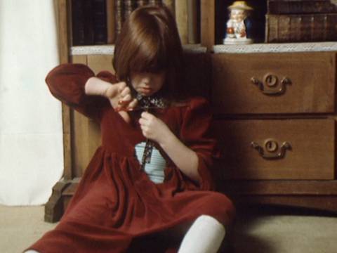 Szenenfoto ›Paulinchen war allein zu Haus‹ BRD 1981, Regie: Anne Voss – Ein kleines Mädchen sitzt vor einem Schrank. Sie trägt ein rotes Kleid, in das sie sich mit einer Schere Löcher schneidet.