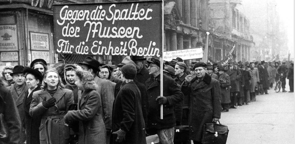Massendemonstration der Berliner Werktätigen, Großkundgebung Unter den Linden zur Konstituierung des neuen Magistrats in der sowjetischen Zone, 30. November 1948 – Schwarzweißfotografie: Menschenmenge auf Straße, Plakattext: "Gegen die Spalter der Muskeln. Für die Einheit Berlins". Erwachsene in Winterkleidung.