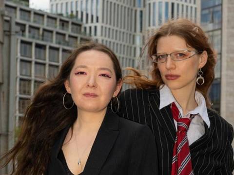  – Zwei Personen in formeller Kleidung vor modernen Hochhäusern; eine trägt eine Brille, die andere große Ohrringe.