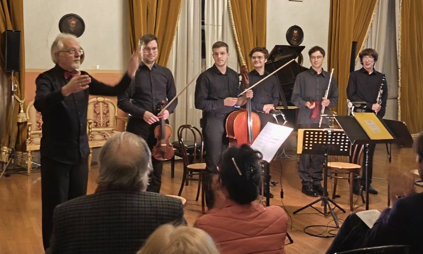 Kammerorchester Hannover-Solisten – Sechs Personen stehen in einem Raum mit gelben Vorhaengen, vier mit Instrumenten. Sitzende Personen im Vordergrund. Ein Dirigent hebt den Arm.