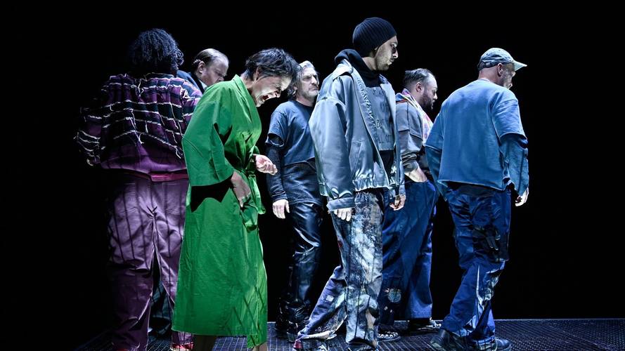 Personengruppe auf einer Bühne, verschiedene bunte Outfits, darunter grüne und blaue Kleidung, auf schwarzem Hintergrund. Keine erkennbare Interaktion.