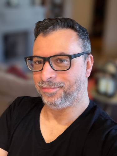 Fabrice Sapolsky – Person mit Brille und Bart, trägt ein schwarzes T-Shirt, sitzt lächelnd. Hintergrund unscharf.