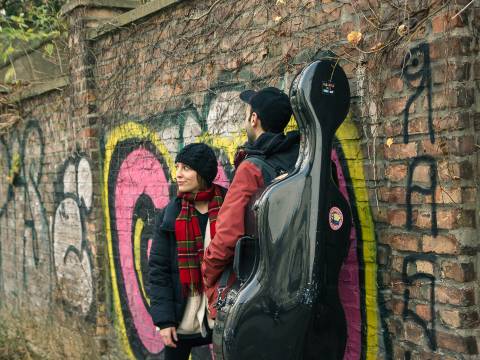  – Zwei Personen stehen vor einer Backsteinwand, die mit Graffiti besprayt ist. Bei der linken Person handelt es sich um die Gastgeberin Antonia Regis. Die rechte Person ist nur von hinten zu sehen, sie trägt ein Cello-Koffer.