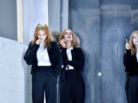  – Drei Personen in schwarz-weißen Outfits mit Zigaretten, an Wand gelehnt, mit blonden Haaren und rosa Make-up, in nachdenklicher Pose.